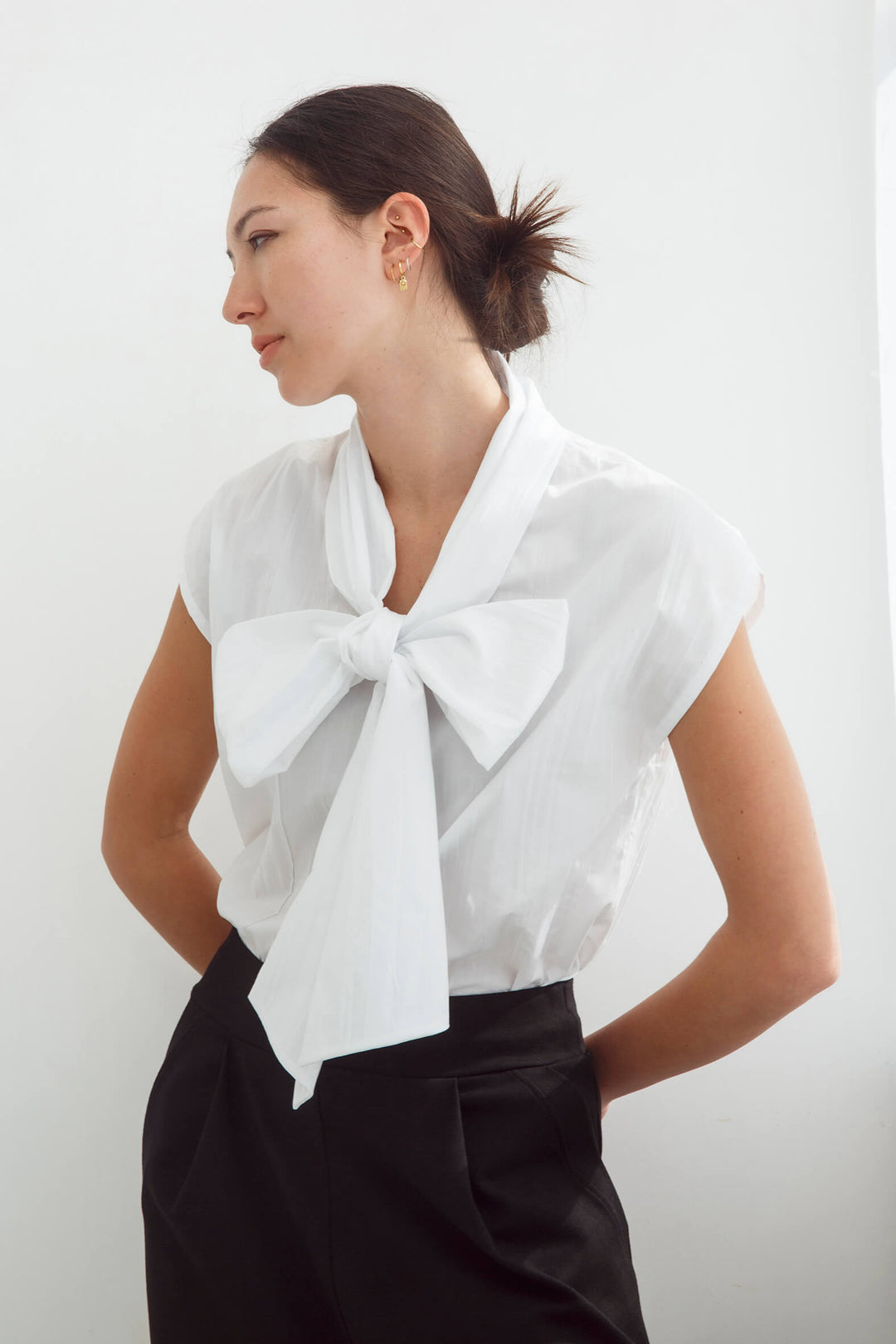 Woman wearing a handmade pleated japanese white sleeveless top with a big bow tie and black pants against a white background