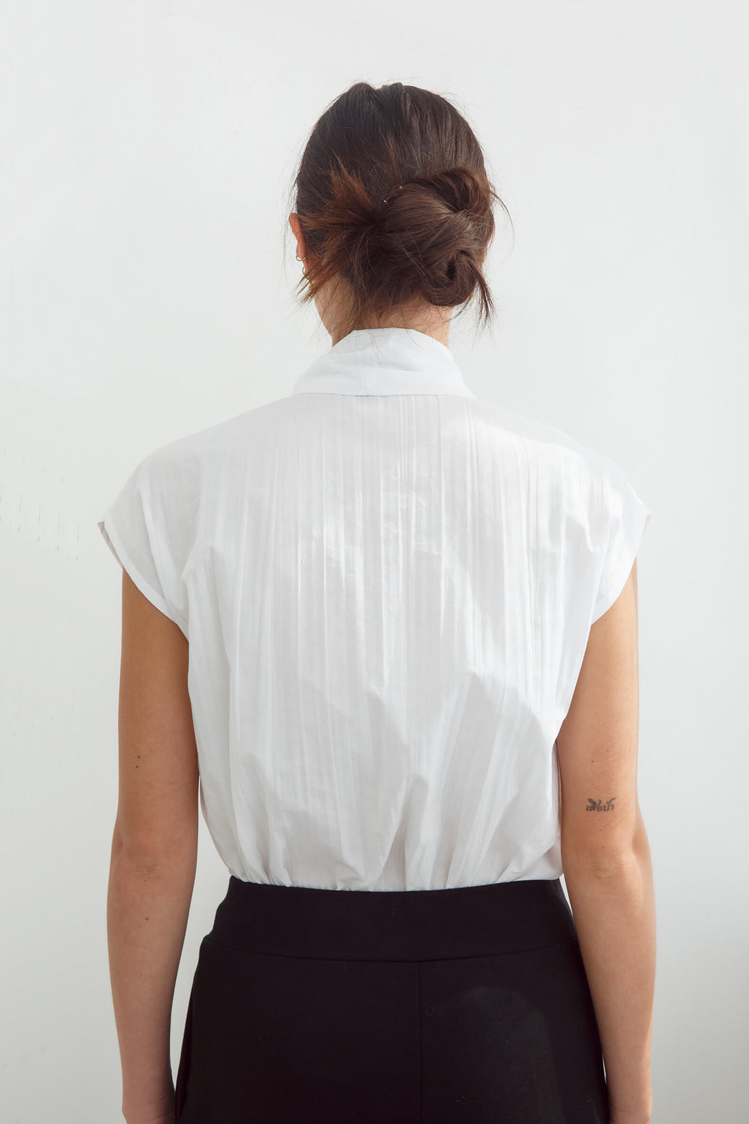 Woman wearing a handmade pleated japanese white sleeveless top with a big bow tie and black pants against a white background