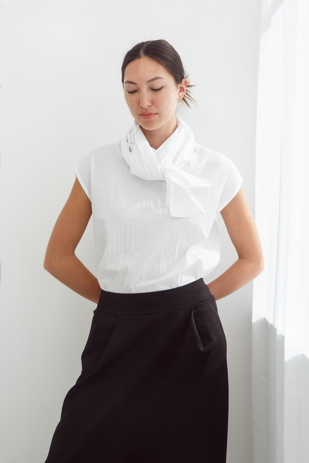 Woman wearing a handmade pleated japanese white sleeveless top with a big bow tie and black pants against a white background