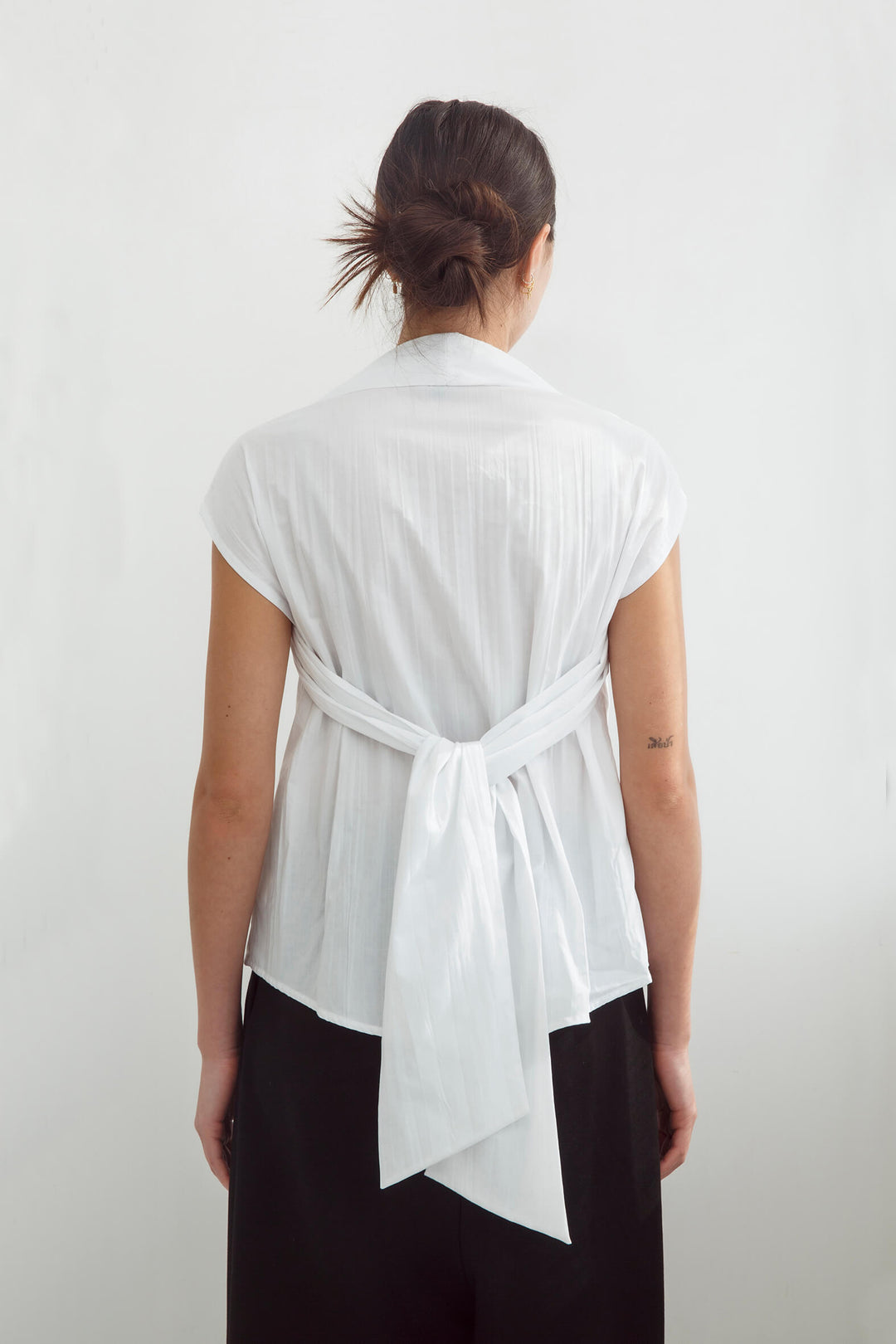 Woman wearing a handmade pleated japanese white sleeveless top with a big bow tie and black pants against a white background