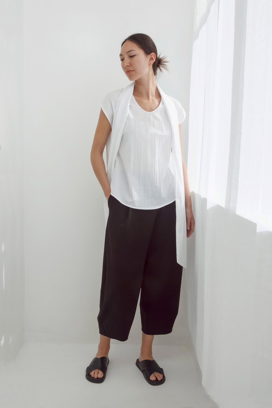 Woman wearing a handmade pleated japanese white sleeveless top with a big bow tie and black pants against a white background