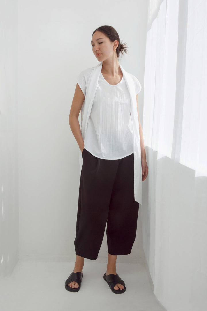 Woman wearing a handmade pleated japanese white sleeveless top with a big bow tie and black pants against a white background
