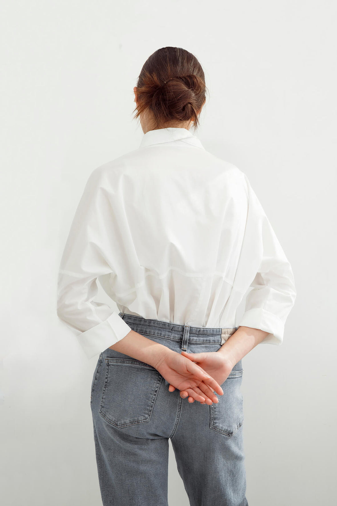 A woman viewed from behind wearing a white long-sleeve button-up shirt tucked into light blue jeans, standing against a plain white wall.