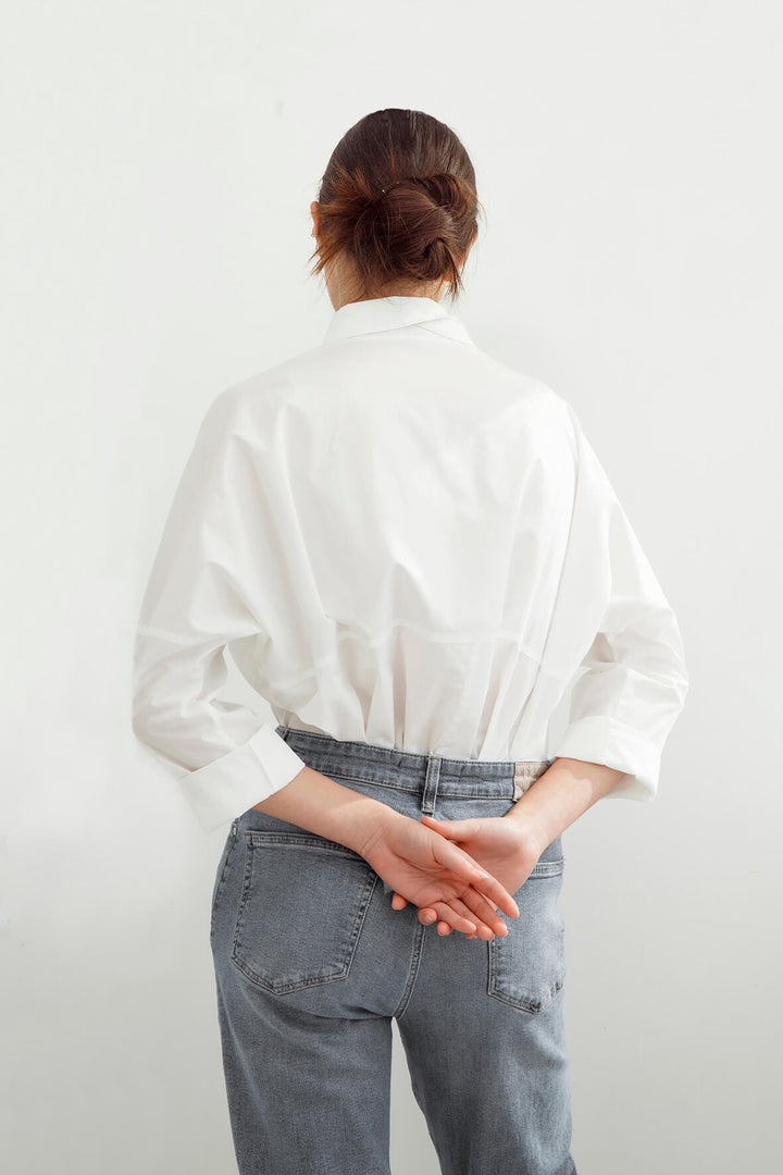 A woman viewed from behind wearing a white long-sleeve button-up shirt tucked into light blue jeans, standing against a plain white wall.