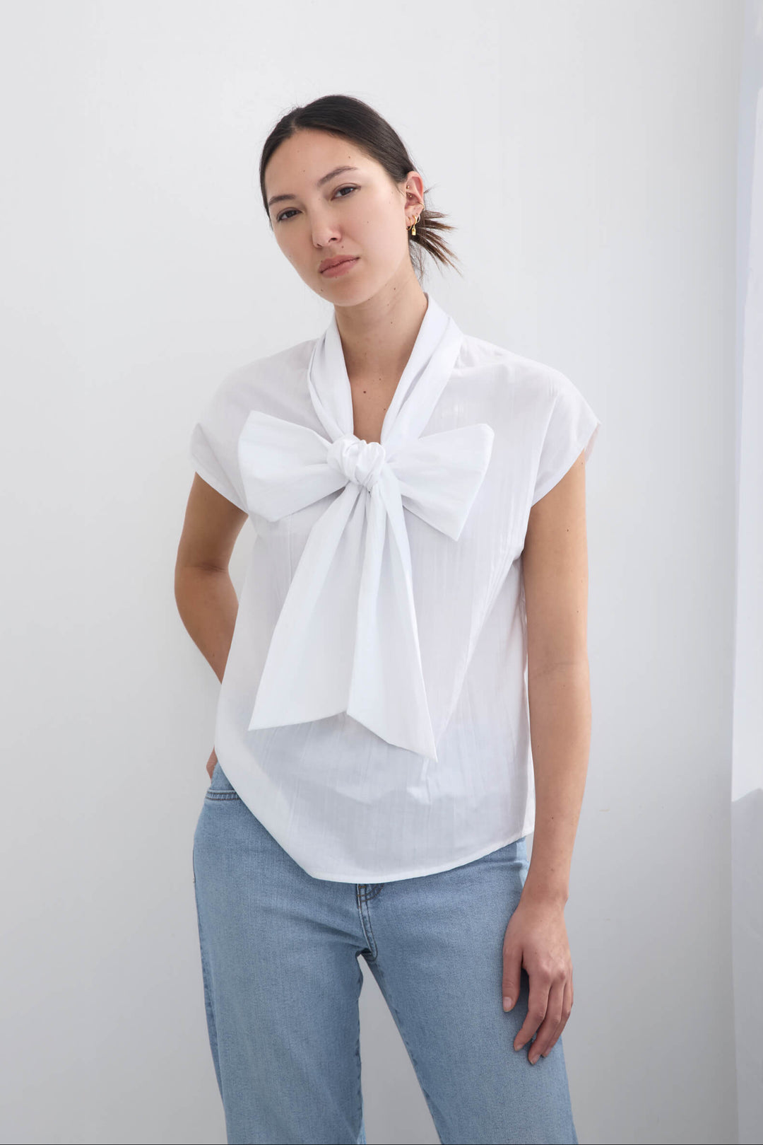 Woman wearing a handmade pleated japanese white sleeveless top with a big bow tie and jeans against a white background